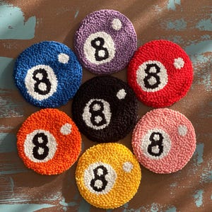 Könnte beinhalten: Sieben bunte, runde Untersetzer, die Billardkugeln mit der Nummer 8 ähneln. Jeder Untersetzer hat eine andere Farbe, darunter Blau, Lila, Rot, Schwarz, Orange, Gelb und Rosa, mit einer weißen 8 und einem weißen Punkt.