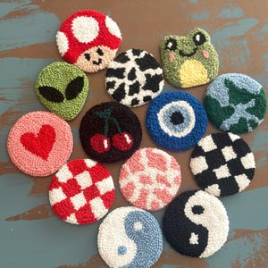 May include: A collection of colorful, round, tufted coasters with various designs. Includes a red mushroom, a green alien, a cow print, a smiling frog, a heart, cherries, an evil eye, a blue and green design, a red and white checkerboard, a pink pattern, a black and white checkerboard, and a yin-yang.