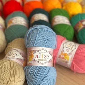 May include: A collection of colorful yarn skeins, including red, orange, green, yellow, and blue. The yarn is labeled "Alize Cotton Gold". The skeins are arranged on a wooden surface, ideal for knitting and crochet projects.