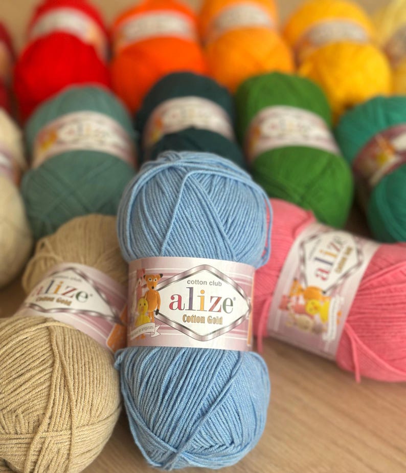 May include: A collection of colorful yarn skeins, including red, orange, green, yellow, and blue. The yarn is labeled "Alize Cotton Gold". The skeins are arranged on a wooden surface, ideal for knitting and crochet projects.