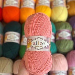May include: A close-up of a ball of Alize Cotton Gold yarn in a soft peach color. The label reads "alize Cotton Gold". Other balls of yarn in various colors, including orange, yellow, green, and purple, are visible in the background.