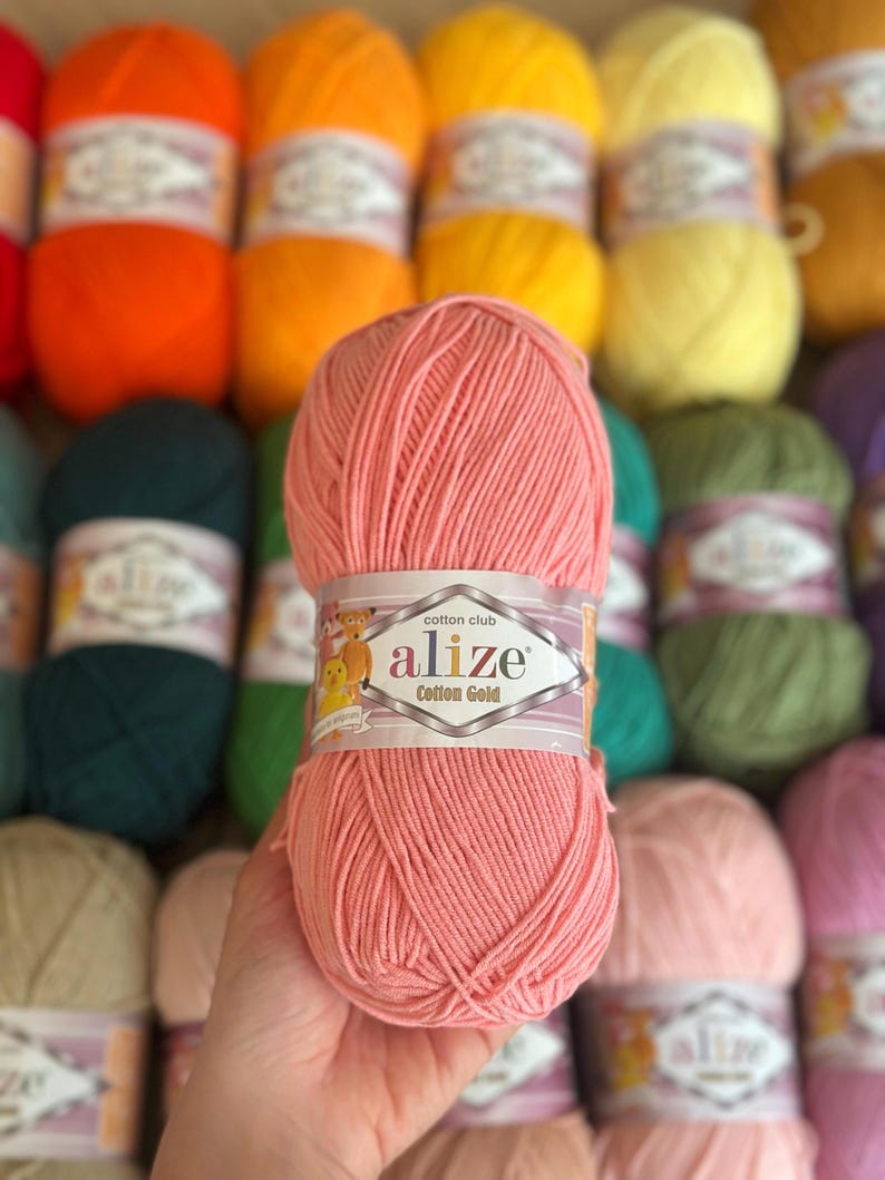 May include: A close-up of a ball of Alize Cotton Gold yarn in a soft peach color. The label reads "alize Cotton Gold". Other balls of yarn in various colors, including orange, yellow, green, and purple, are visible in the background.