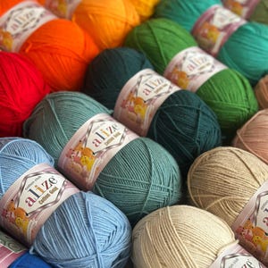 May include: A collection of colorful yarn balls in shades of red, orange, yellow, green, blue, and beige. Each ball is labeled with the brand "Alize Cotton Gold". The yarn is arranged in rows, creating a visually appealing display for crafting projects.