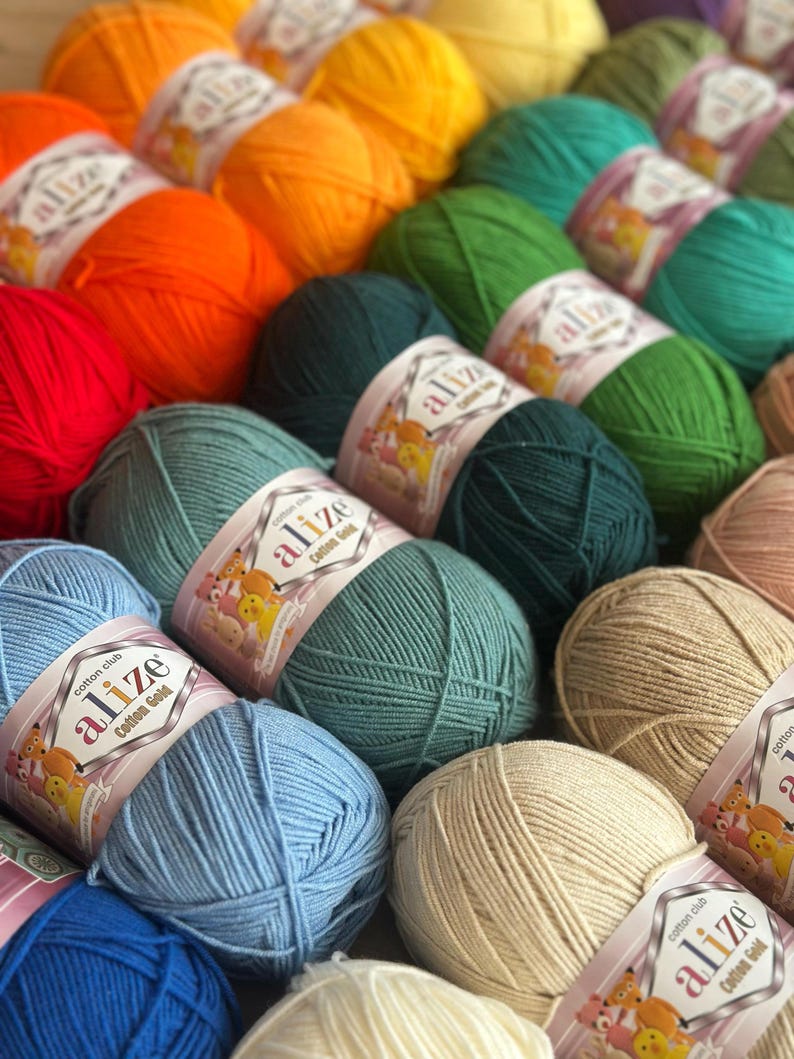 May include: A collection of colorful yarn balls in shades of red, orange, yellow, green, blue, and beige. Each ball is labeled with the brand "Alize Cotton Gold". The yarn is arranged in rows, creating a visually appealing display for crafting projects.