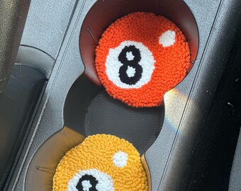 Può includere: Due sottobicchieri rotondi per auto, uno arancione e uno giallo, ciascuno con un design a palla numero 8 bianca. I sottobicchieri sono posizionati nei portabicchieri di un'auto, offrendo un accessorio divertente e funzionale.