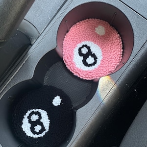 May include: Two handmade car coasters, one pink and one black, each featuring a crocheted design of an eight ball. The pink coaster has a white dot and black number eight. The black coaster has a white dot and a white number eight.
