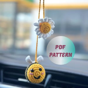 Op de afbeelding: Een handgemaakte gehaakte auto-hanger met een lachende gele bij met zwarte strepen en witte vleugels, en een madeliefje met witte bloemblaadjes en een geel hart. De hanger hangt aan een gele draad. Tekst op de afbeelding zegt "PDF PATTERN".