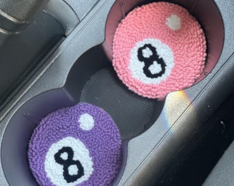 Può includere: Due sottobicchieri per auto, uno rosa e uno viola, ciascuno simile a una palla numero otto. I sottobicchieri sono rotondi con un cerchio bianco e il numero 8 al centro. I sottobicchieri sono nei portabicchieri di un'auto.