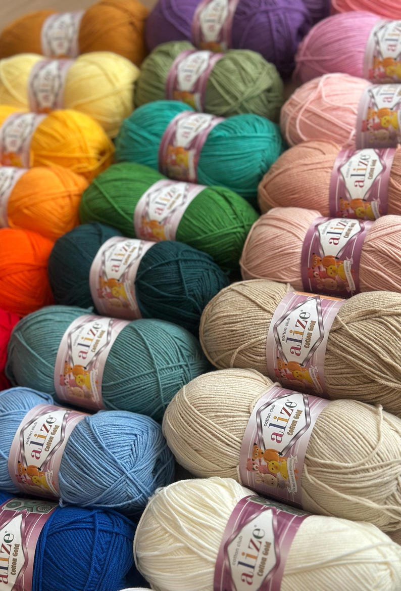 May include: A collection of colorful yarn skeins in various shades, including yellow, green, pink, and blue. Each skein is labeled with the brand name "alize Cotton Gold". The yarn is neatly arranged, suggesting a craft supply or textile product.