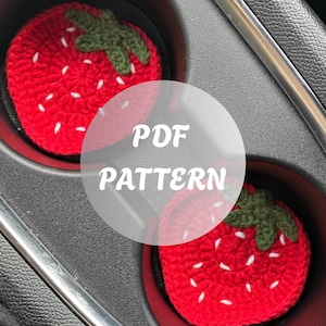 May include: Two red crochet strawberry car coasters with white seed details and green leaves. The coasters are in a car cup holder. The text "PDF PATTERN" is overlaid on the image. The text "Loops by Angelina" is in the bottom left corner.