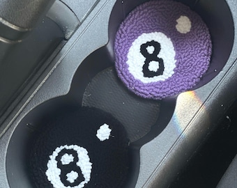 Può includere: Due sottobicchieri per auto, uno nero e uno viola, ciascuno con un numero 8 bianco e un punto bianco, che assomigliano alla palla 8 del biliardo. I sottobicchieri sono nei portabicchieri di un'auto.