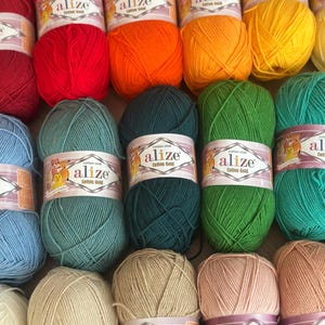 May include: A collection of colorful Alize Cotton Gold yarn skeins. The yarn is available in a variety of colors, including red, orange, yellow, green, blue, beige, and pink. Each skein is labeled with the "Alize Cotton Gold" brand name.