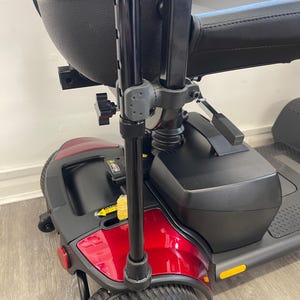 May include: A red and black mobility scooter with a black seat and armrest. The scooter has a black frame and a front wheel with a silver hubcap. The scooter has a black tiller and a black storage compartment.