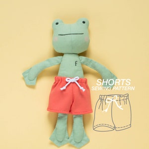 May include: A green frog plush toy wearing red shorts with a white drawstring. The image also includes a line drawing of a pair of shorts with a drawstring, labeled "Shorts Sewing Pattern".