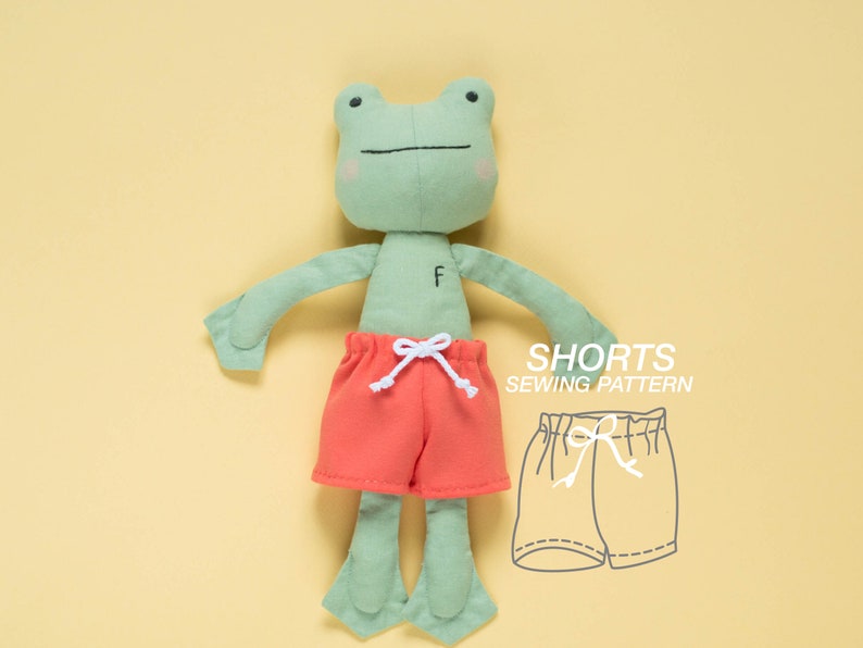May include: A green frog plush toy wearing red shorts with a white drawstring. The image also includes a line drawing of a pair of shorts with a drawstring, labeled "Shorts Sewing Pattern".