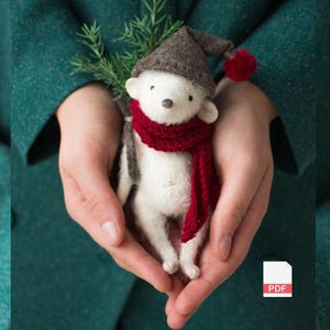 May include: A handmade white felt animal figurine with a red scarf and a brown hat with a red pom-pom. The figurine is holding a small sprig of greenery. The figurine is held in cupped hands.
