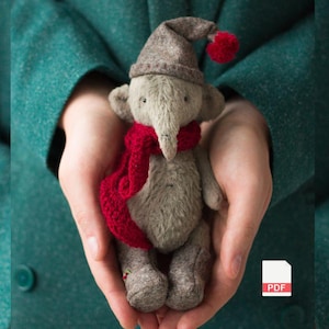 May include: A handmade gray elephant plush toy wearing a brown hat with red pom-poms and a red scarf. The elephant is held in cupped hands against a teal background. The toy has a long trunk and large ears.