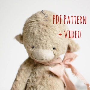 May include: A handmade, plush teddy bear with a light brown fur texture, small black eyes, and a stitched nose. It has a pink ribbon around its neck. The text "PDF PATTERN + VIDEO" is written in red.