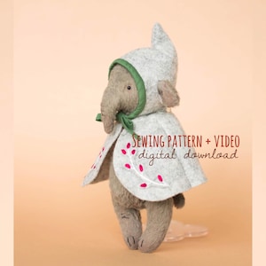 Könnte beinhalten: Ein Plüschelefant trägt einen hellgrauen Kapuzenumhang mit grünem Saum und rosa Blumenstickerei. Der Text SEWING PATTERN + VIDEO digital download ist in Rot und Schwarz auf der rechten Seite der Elefantenfigur aufgedruckt.