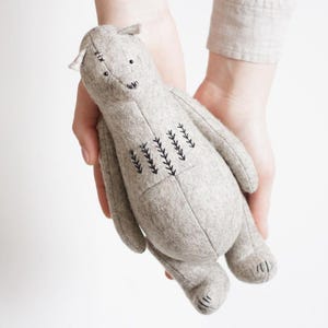 Peut inclure: Une peluche en tissu feutré gris clair en forme d'ours. Le jouet présente des détails brodés au fil foncé sur le visage, les oreilles et la poitrine, avec de petits yeux en perles noires et des griffes cousues sur les pattes, avec des coutures visibles.