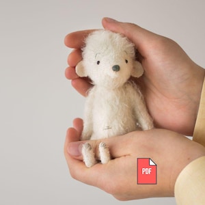 Puede incluir: Un pequeño oso de peluche blanco hecho a mano con una nariz gris claro y ojos negros pequeños. El oso se sostiene con dos manos. El oso tiene orejas y pies pequeños. Un icono PDF está en la esquina inferior derecha.