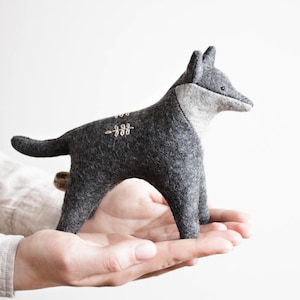 May include: A small, handcrafted felt animal figurine in the shape of a fox or wolf. The body is made of dark charcoal gray felt with a lighter gray chest and face. It features simple white embroidered leaf details on its back and a small tag.