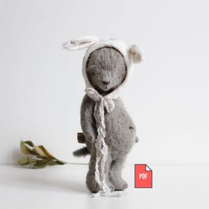 Può includere: Un orsacchiotto grigio fatto a mano con un cappuccio bianco con orecchie flosce e una cravatta abbinata. L'orso ha una piccola coda ed è in piedi. Un'icona PDF si trova nell'angolo in basso a destra.