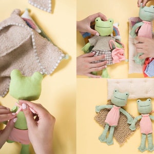 May include: Two green frog plushies with embroidered faces and pink dresses. One frog is lying on a tan blanket with a white pillow. The other frog is being dressed by a hand.