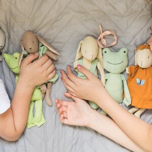 Two children lying on a quilted bedspread, playing with handmade soft dolls dressed in simple fabric outfits, showing scale and play use.