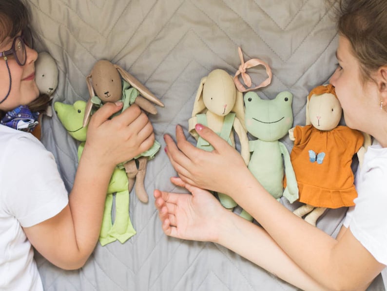 Two children lying on a quilted bedspread, playing with handmade soft dolls dressed in simple fabric outfits, showing scale and play use.