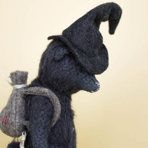 May include: A dark charcoal gray felted animal figurine wearing a matching pointed hat. The figure has a textured, fuzzy surface and carries a small, light gray felted backpack with a red embroidered detail on the back, secured with white string.