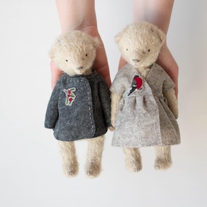 May include: Two beige plush teddy bears with stitched facial features. The left bear wears a dark gray jacket with a small embroidered sprig. The right bear wears a light gray wrap-style dress featuring an embroidered bird with a red chest.