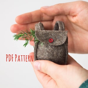 May include: A small brown felt backpack with a single red button closure on the front flap. A sprig of green pine needles is tucked into the side of the bag. The text PDF PATTERN is printed in red on the left side of the image.