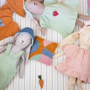 Fabric dolls dressed in handmade outfits lying on striped bedding, showing a variety of doll clothes including dresses, overalls, and textured knitted garments.