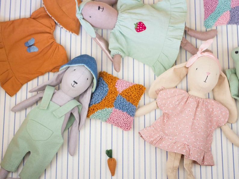 Fabric dolls dressed in handmade outfits lying on striped bedding, showing a variety of doll clothes including dresses, overalls, and textured knitted garments.
