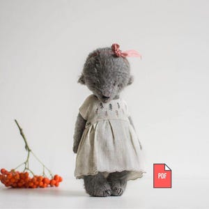 Op de afbeelding: Een grijze teddybeerpop met een licht linnen jurkje en een kleine roze strik. De jurk heeft een geborduurd ontwerp. Een takje met oranje bessen staat op de voorgrond.