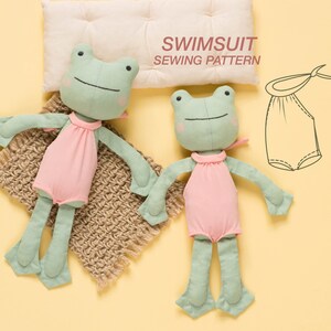 May include: Two green frog plushies wearing pink swimsuits. The plushies are lying on a brown knitted blanket and a white pillow. The pillow has the text "SWIMSUIT SEWING PATTERN" printed on it. A line drawing of a swimsuit pattern is also shown.