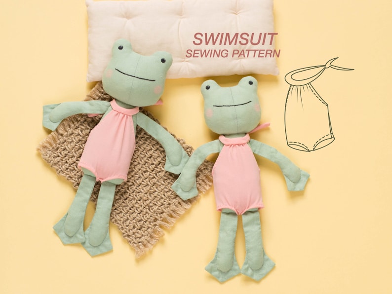 May include: Two green frog plushies wearing pink swimsuits. The plushies are lying on a brown knitted blanket and a white pillow. The pillow has the text "SWIMSUIT SEWING PATTERN" printed on it. A line drawing of a swimsuit pattern is also shown.