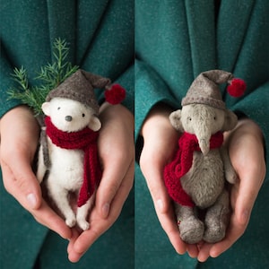 May include: Two handmade plush animal figures, one white and one gray, each wearing a red scarf and a brown hat with a red pom-pom. The white figure holds a sprig of greenery. The figures are held in cupped hands.