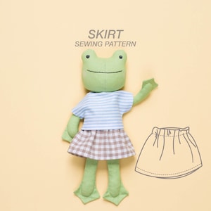 May include: A green frog plush toy wearing a blue and white striped shirt and a brown and white plaid skirt. The image also includes a line drawing of a skirt sewing pattern.