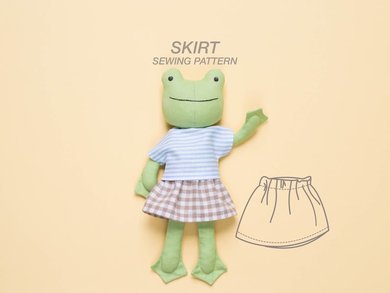 May include: A green frog plush toy wearing a blue and white striped shirt and a brown and white plaid skirt. The image also includes a line drawing of a skirt sewing pattern.
