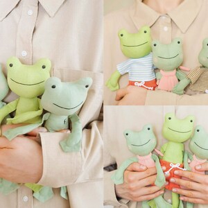 May include: Three green frog plush toys with embroidered faces and different outfits. The frogs are being held by a person's hands.