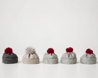 May include: Five small knit hats arranged in a row. The first hat is dark gray with a red pom-pom. The second is light beige with a matching beige pom-pom. The third, fourth, and fifth hats are light gray, each topped with a red pom-pom.