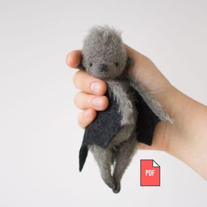 May include: A handmade gray felt bat doll with black felt wings. The doll has a small, detailed face and is held in a hand. The doll is a unique, handcrafted item, perfect for collectors or as a decorative piece. The PDF icon is in the lower right corner.