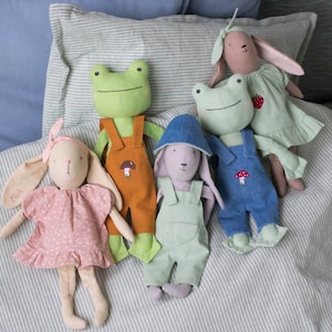 Group of handmade fabric dolls sitting together on a bed, dressed in overalls and dresses, styled as a cozy nursery or playroom scene.