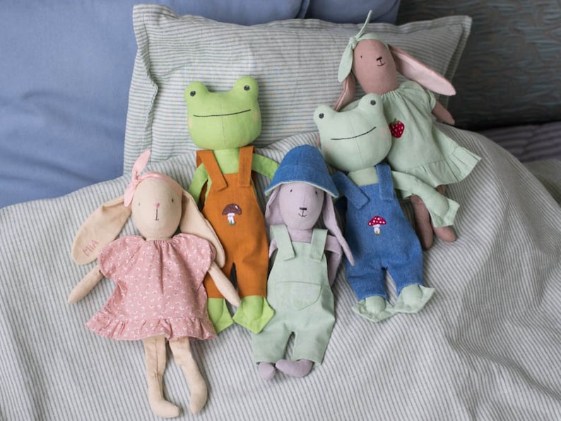 Group of handmade fabric dolls sitting together on a bed, dressed in overalls and dresses, styled as a cozy nursery or playroom scene.