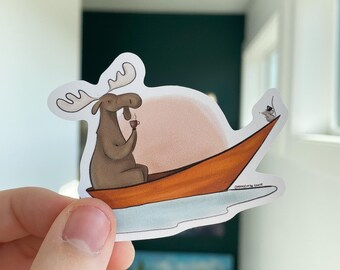 May include: A brown moose with large antlers sits in an orange canoe, holding a cup of coffee. The canoe is floating on a blue lake with a pink sunset in the background. The text "Inspired by the Coast" is printed on the bottom of the canoe.
