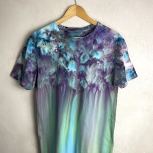 Könnte beinhalten: Kurzärmeliges T-Shirt mit Batikmuster in Blau-, Lila- und Grüntönen. Die Farben verschmelzen in einem vertikalen Farbverlauf und erzeugen ein einzigartiges und künstlerisches Design. Das T-Shirt hängt an einem Holzkleiderbügel.