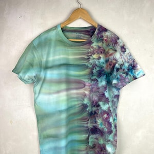 Könnte beinhalten: Ein kurzärmeliges T-Shirt mit einem einzigartigen Batik-Design. Die linke Seite zeigt horizontale Streifen in Blau-, Grün- und Lilatönen. Die rechte Seite hat ein Blumenmuster in Lila, Blau und Grün. Das Shirt hängt an einem Holzbügel.