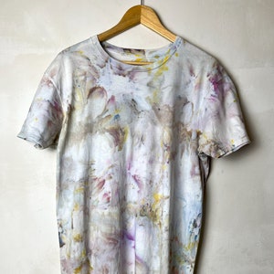 May include: A white t-shirt with a tie-dye pattern in shades of pink, yellow, and blue. The shirt is hanging on a wooden hanger.